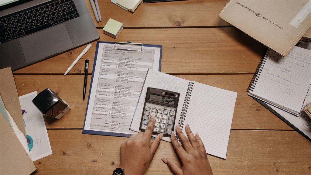 Mesa com calculadora, caneta e papéis de cálculo ao lado de um notebook
