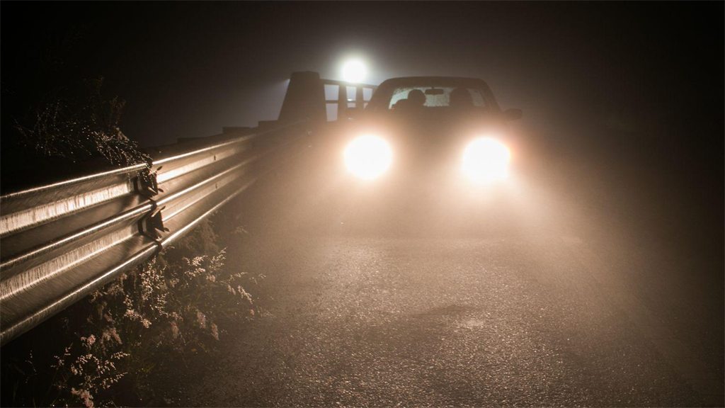 Faróis de um carro acesos em estrada com neblina densa